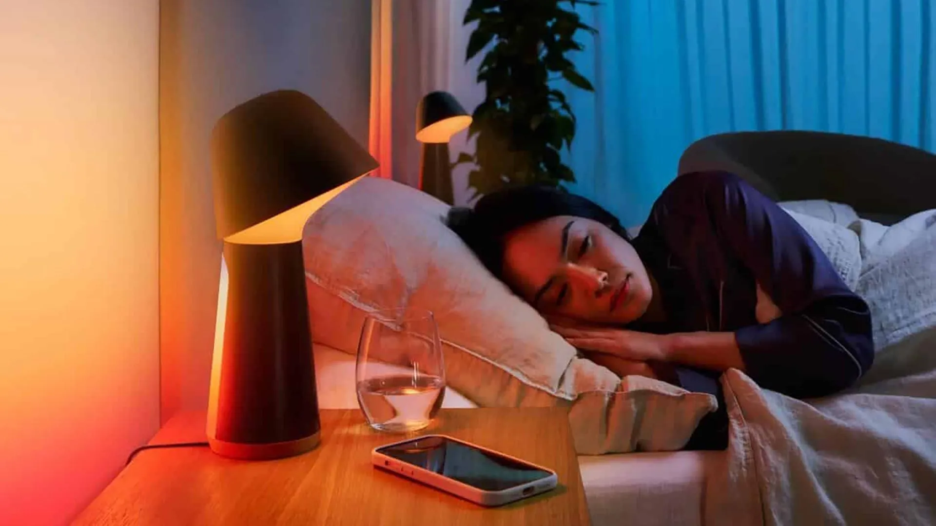 Người phụ nữ đang ngủ sâu giấc trên giường, cạnh đó là đèn ngủ thông minh và điện thoại thông minh, thể hiện môi trường nghỉ ngơi lý tưởng.
