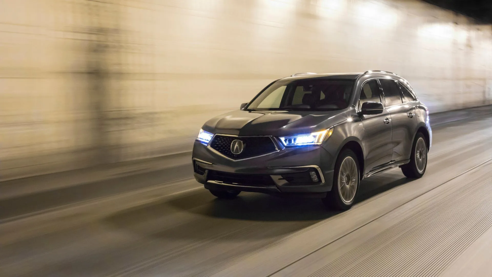Acura MDX Sport Hybrid 2020 màu xám di chuyển qua cầu vượt với đèn pha bật, thể hiện tầm quan trọng của hệ thống chiếu sáng an toàn trên đường