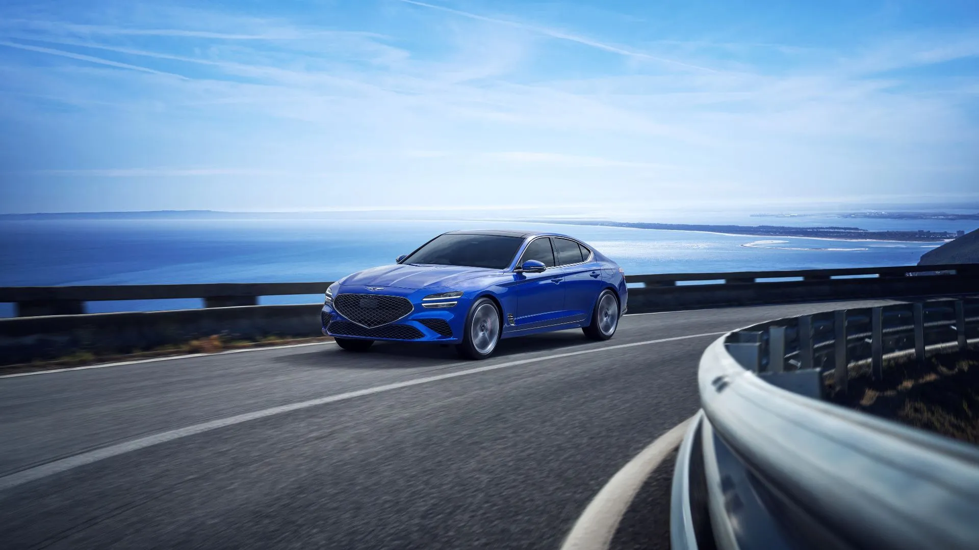 Chiếc Genesis G70 2025 màu xanh đang lướt đi trên con đường ven biển uốn lượn, cảnh quan đại dương tuyệt đẹp phía sau, toát lên vẻ đẳng cấp và phong cách sống thượng lưu.