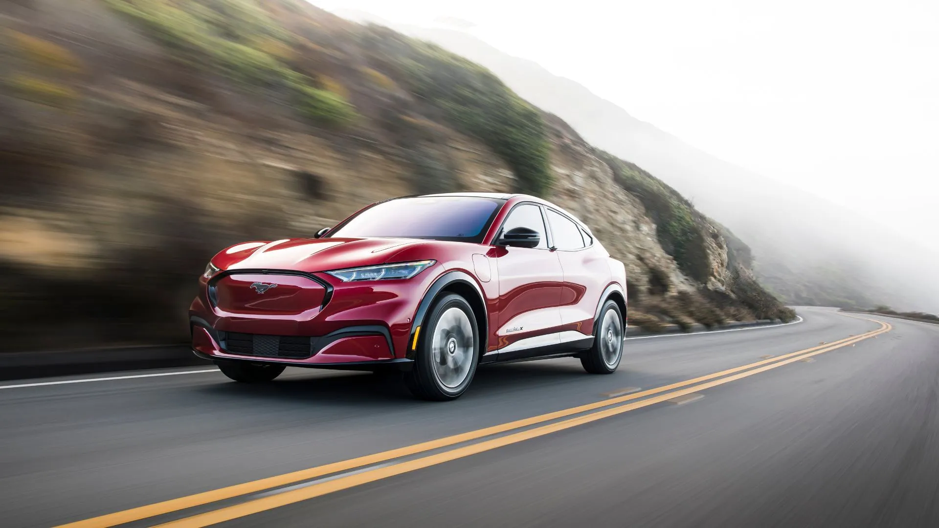 Ford Mustang Mach-E 2025 màu đỏ, toàn cảnh, đang lái trên con đường nông thôn quanh co, biểu tượng cho hiệu suất xe điện.