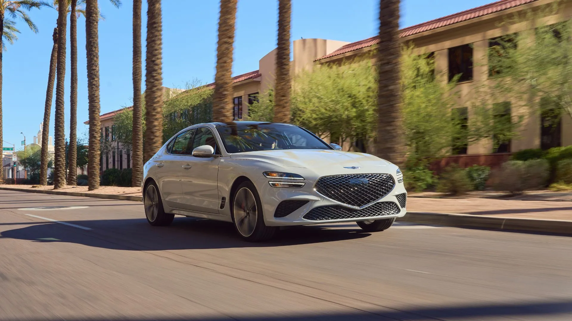 Genesis G70 2025 màu xám bạc đang di chuyển qua khu phố đô thị sầm uất với hàng cọ xanh mát hai bên, thể hiện sự phù hợp của mẫu xe sang này trong mọi bối cảnh, từ đường trường đến thành phố.