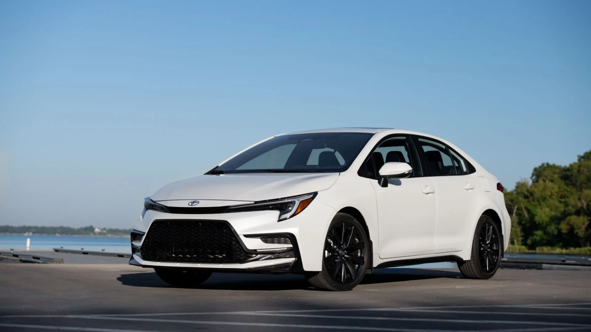 Góc 3/4 phía trước của Toyota Corolla Sedan 2025 màu trắng, đậu với bối cảnh biển xanh, thể hiện sự đơn giản và hiệu quả nhiên liệu.