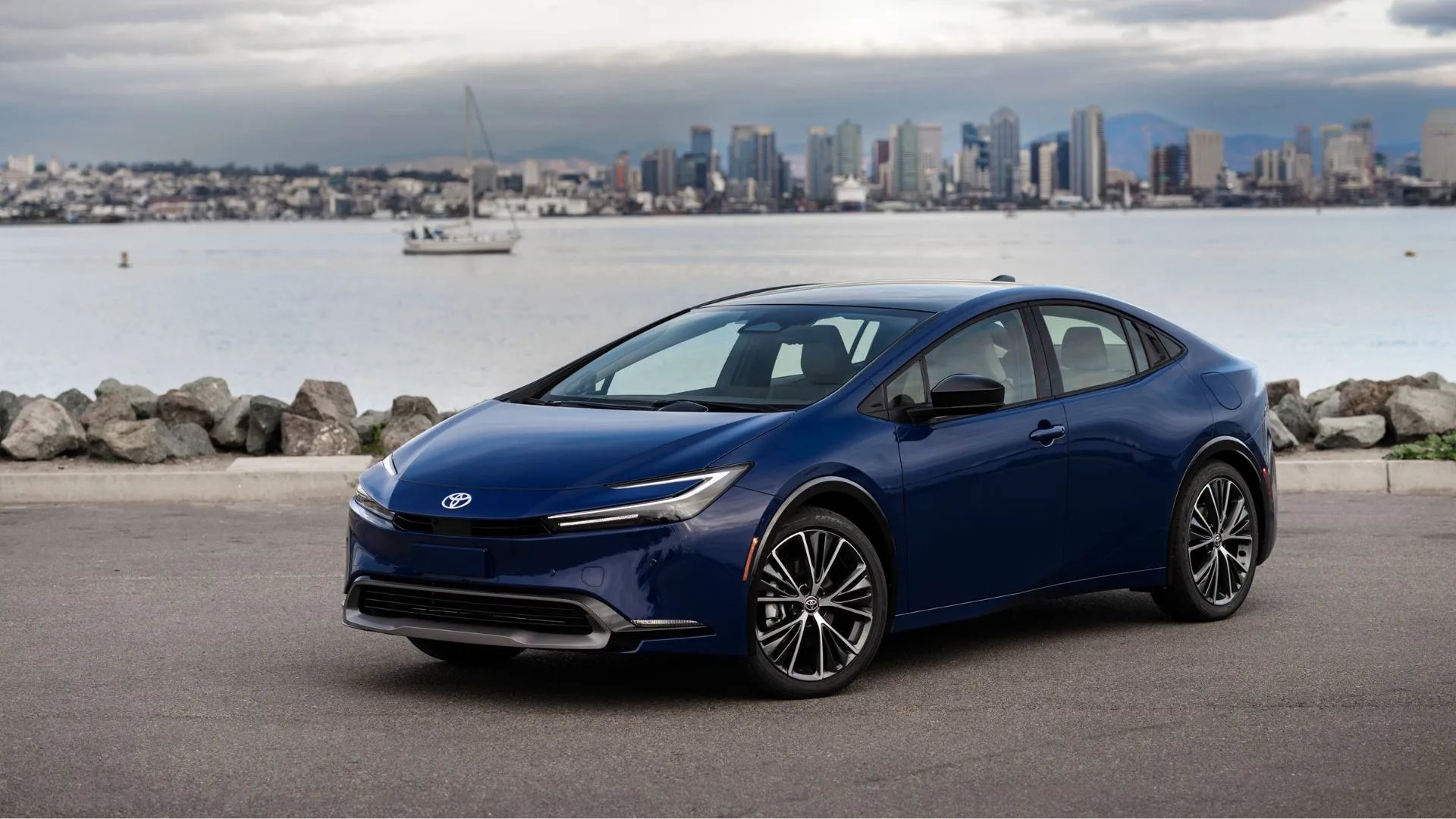 Góc 3/4 phía trước của Toyota Prius 2025 màu xanh đậm, với cảnh quan mặt nước và đường chân trời thành phố, khẳng định vị thế dẫn đầu về tiết kiệm nhiên liệu.