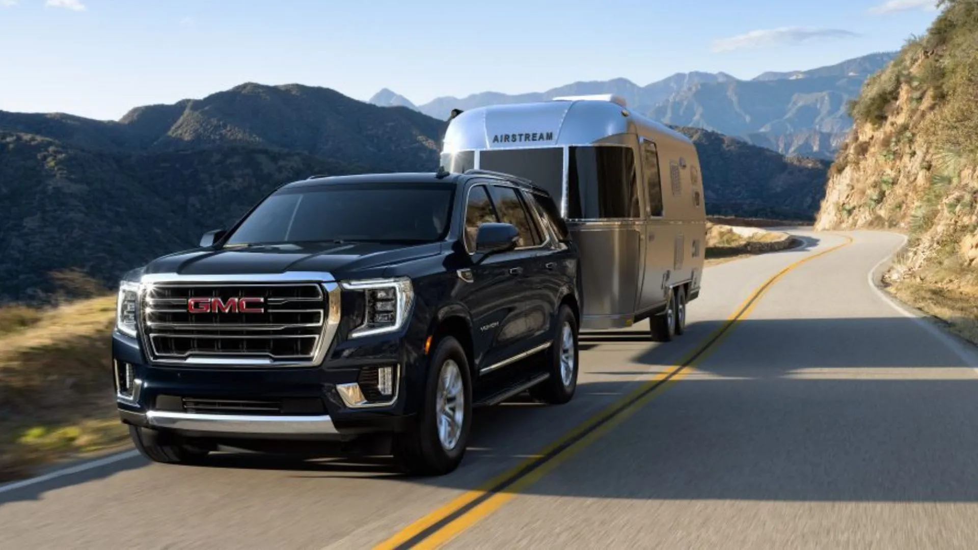 Góc ba phần tư phía trước của chiếc GMC Yukon 2022 màu đen đang kéo rơ-moóc trên đường nông thôn qua núi.