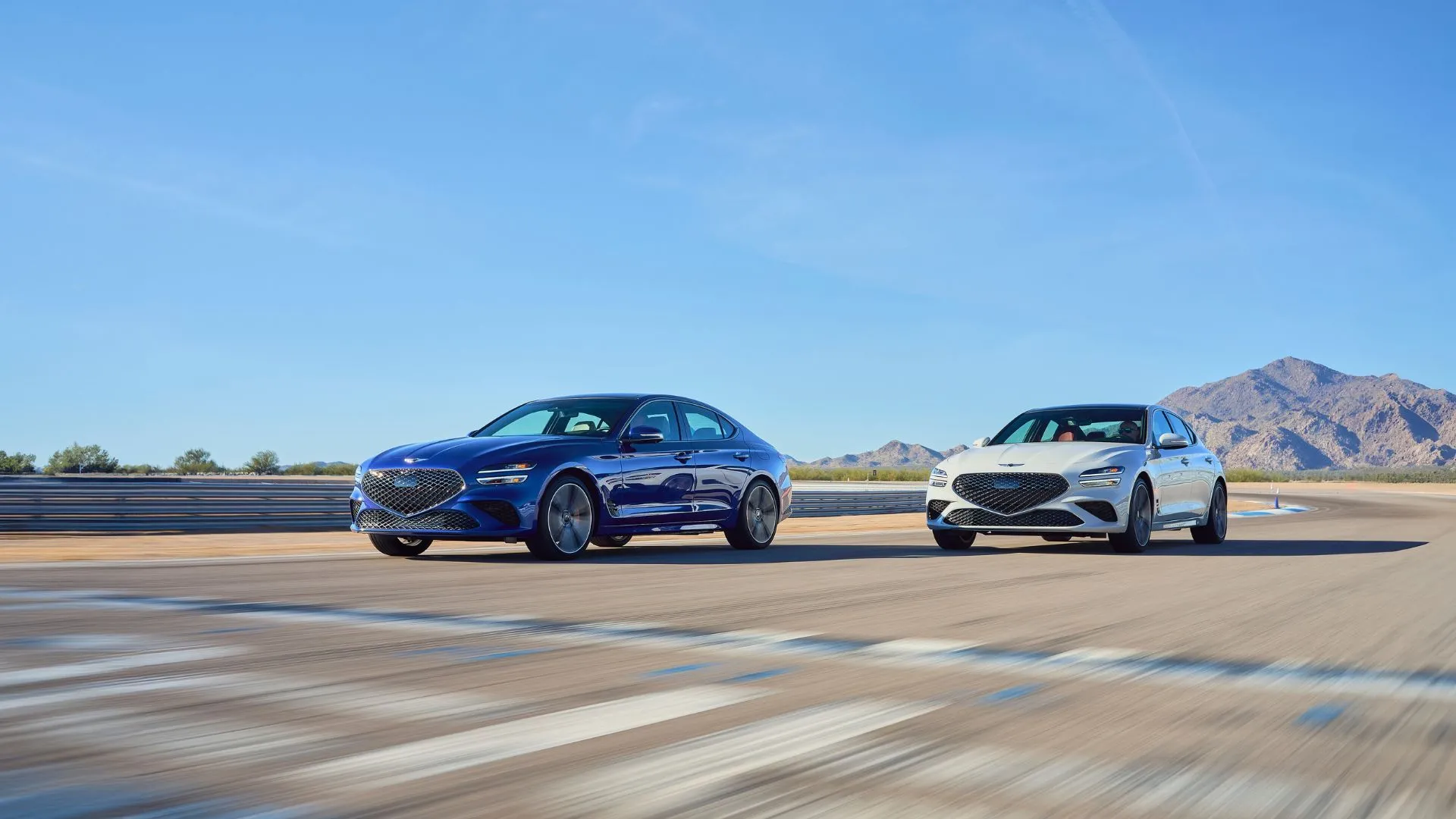 Hai chiếc Genesis G70 2025 màu xanh và xám đang tăng tốc mạnh mẽ trên đường đua dragstrip, minh họa rõ nét khả năng vận hành thể thao và hiệu suất động cơ vượt trội của mẫu sedan này.