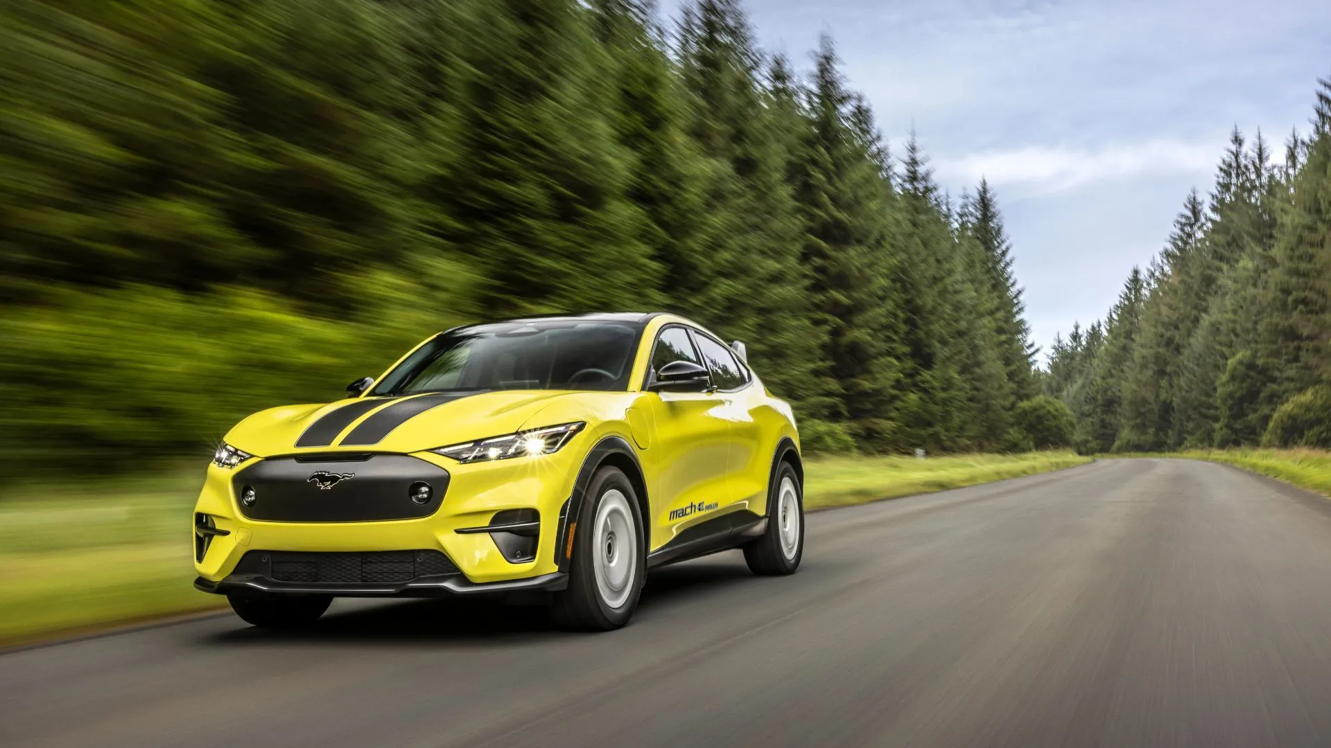 Hình ảnh mặt trước ba phần tư năng động của một chiếc Ford Mustang Mach-E Rally màu vàng đang lái trên đường sỏi xuyên rừng.