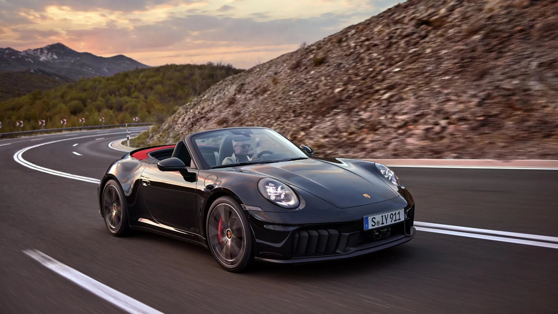 Hình ảnh toàn cảnh Porsche 911 Carrera GTS Cabriolet Hybrid 2024 màu đen đang chạy trên con đường nông thôn quanh co với núi non phía sau