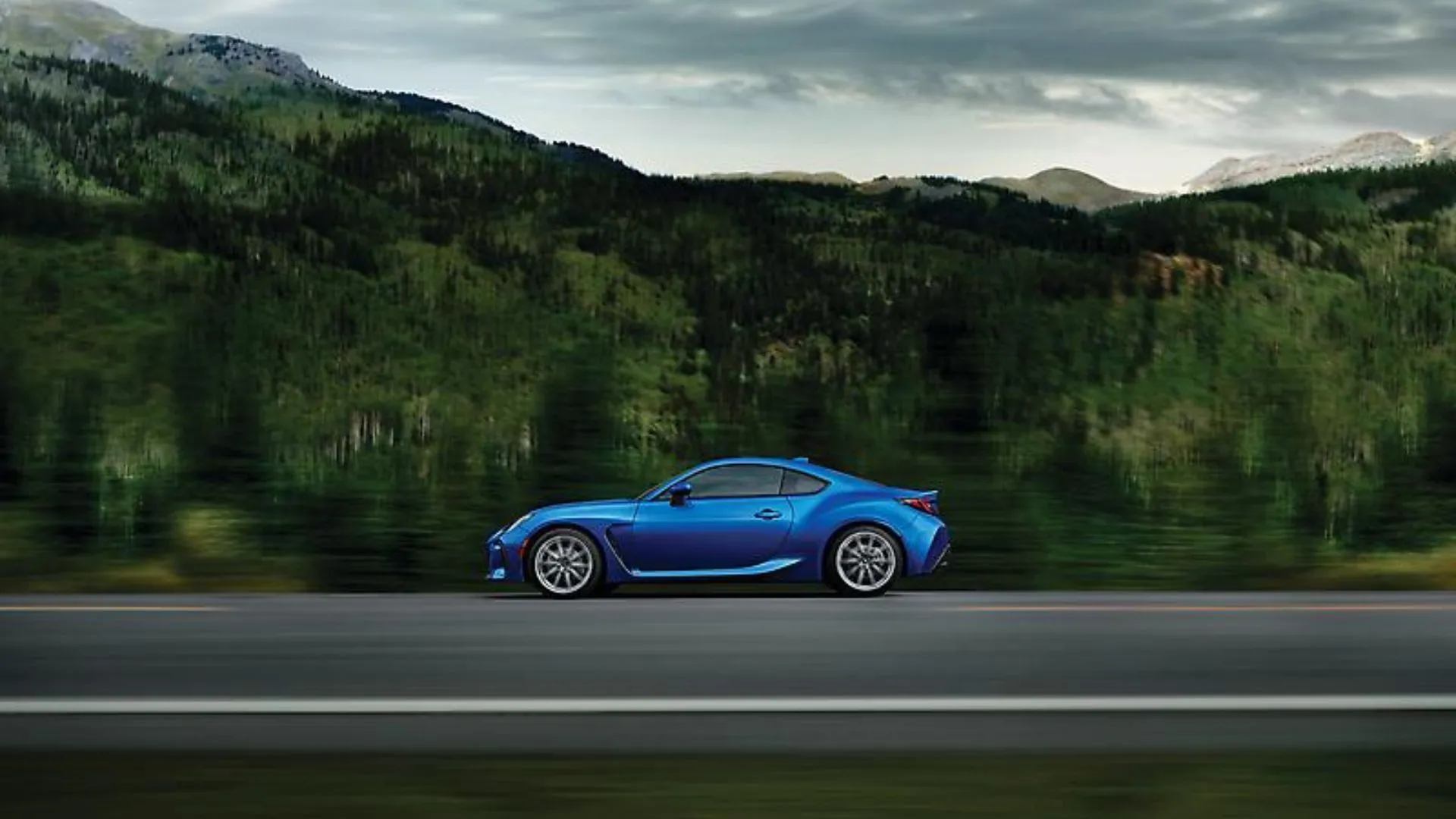 Hồ sơ bên hông năng động của Subaru BRZ 2024 màu xanh chạy trên con đường nông thôn, với núi non và cây cối làm nền