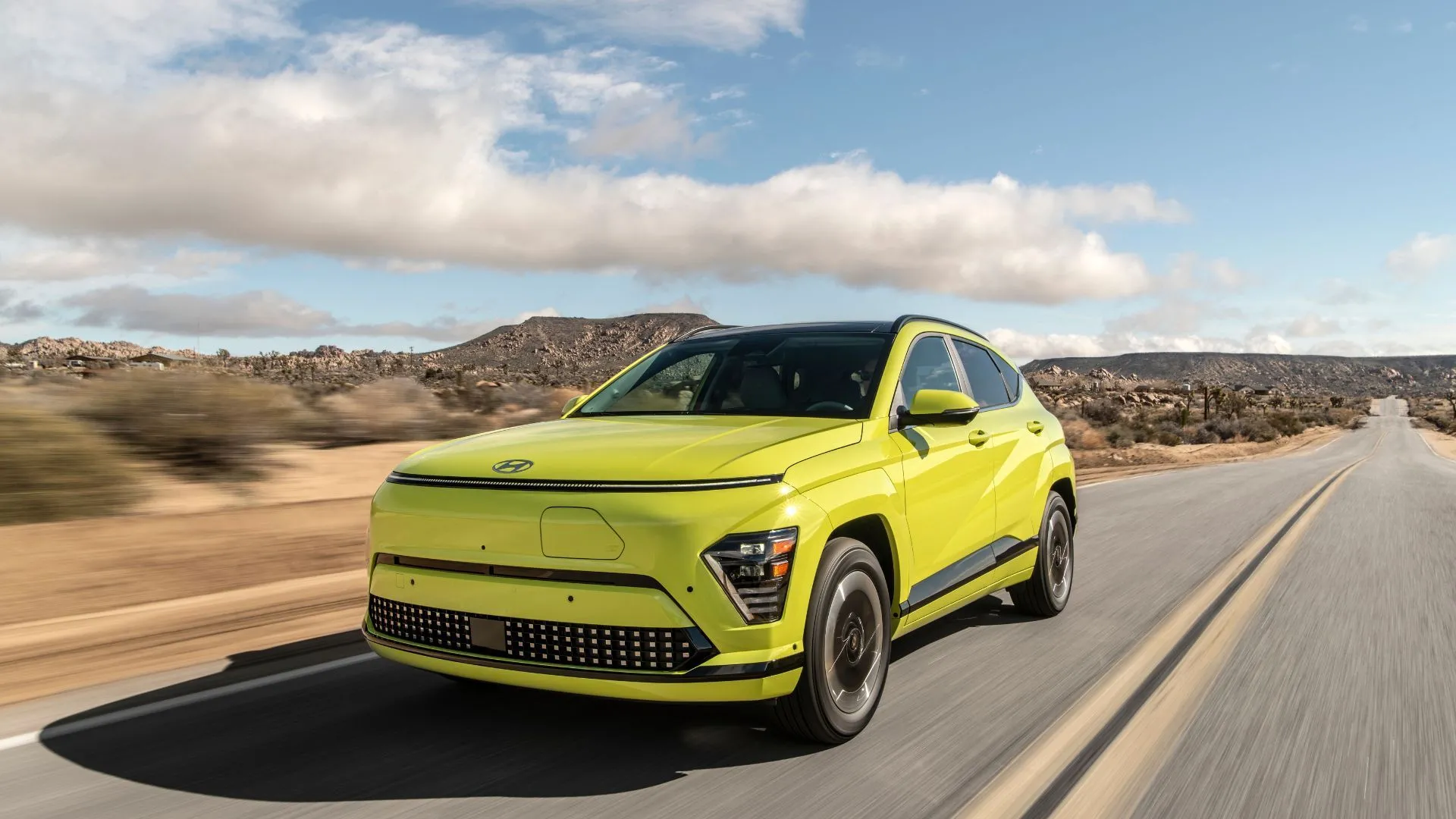 Hyundai Kona Electric 2025 màu vàng, góc 3/4 phía trước, đang di chuyển trên đường sa mạc, thể hiện sự linh hoạt của crossover điện.