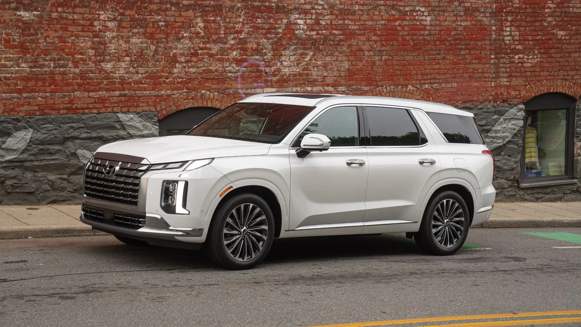 Hyundai Palisade 2025 màu trắng nhìn từ góc 3/4 phía trước đỗ bên vỉa hè với tường gạch graffiti