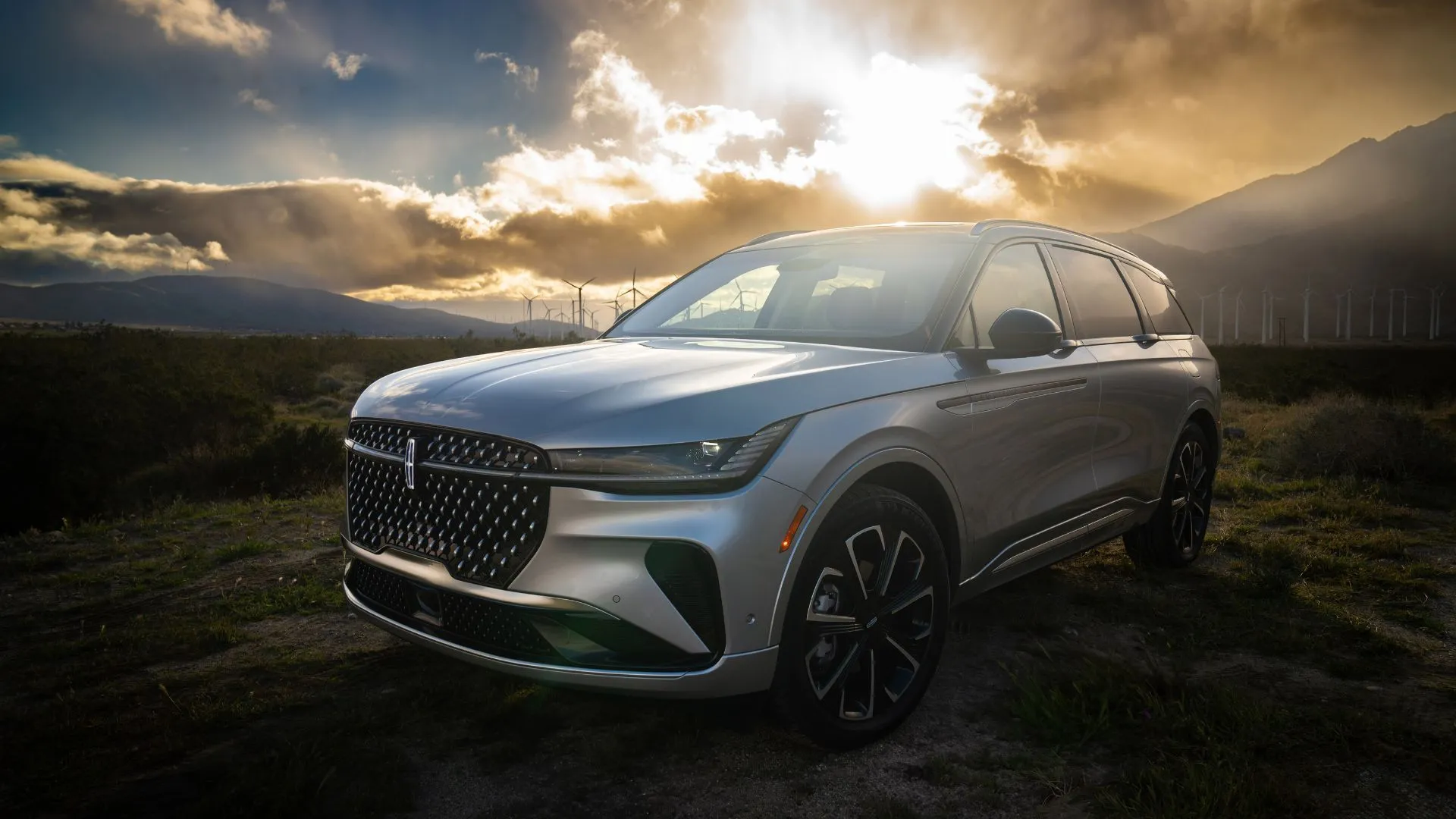 Lincoln Nautilus Reserve Hybrid 2024 màu bạc đỗ vào lúc hoàng hôn, với các tuabin gió ở phía sau, biểu thị sự kết hợp giữa sang trọng và bền vững