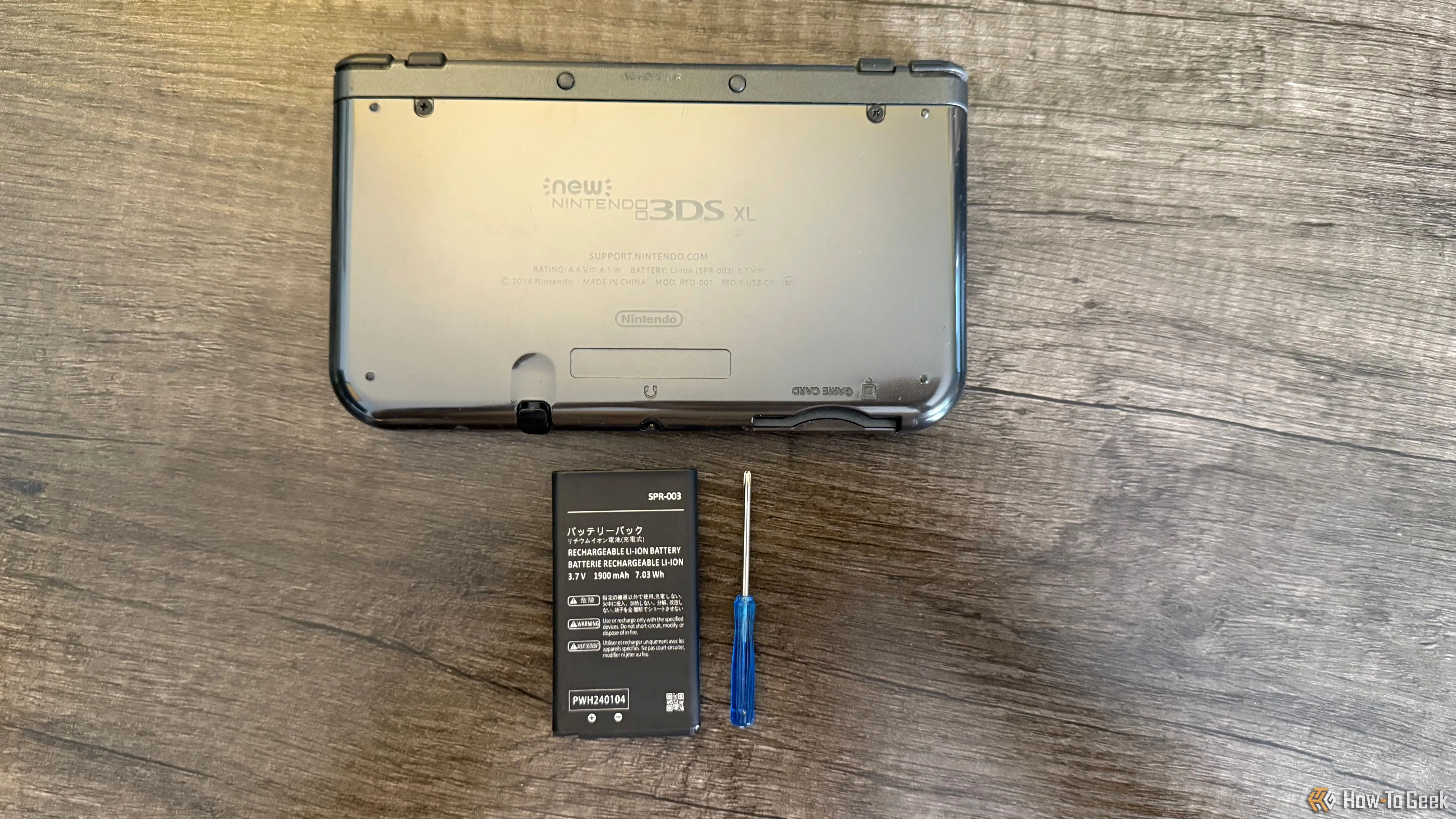 Mặt sau của máy chơi game New Nintendo 3DS XL màu đen, bên cạnh là pin thay thế và một tua vít đầu Phillips nhỏ, chuẩn bị tháo nắp.