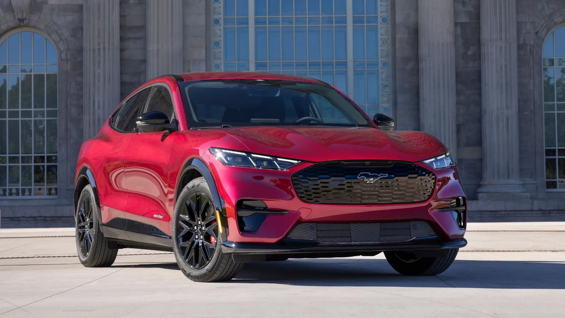 Mặt trước 3/4 của chiếc Ford Mustang Mach-E 2025 màu đỏ với một tòa nhà ở hậu cảnh.