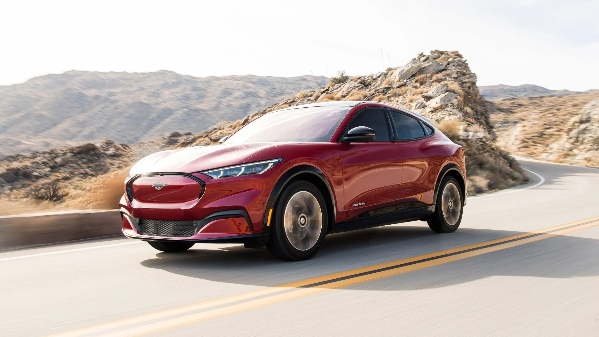 Mặt trước 3/4 của chiếc Mustang Mach-E 2021 màu đỏ đang lái trên đường nông thôn với núi ở hậu cảnh.