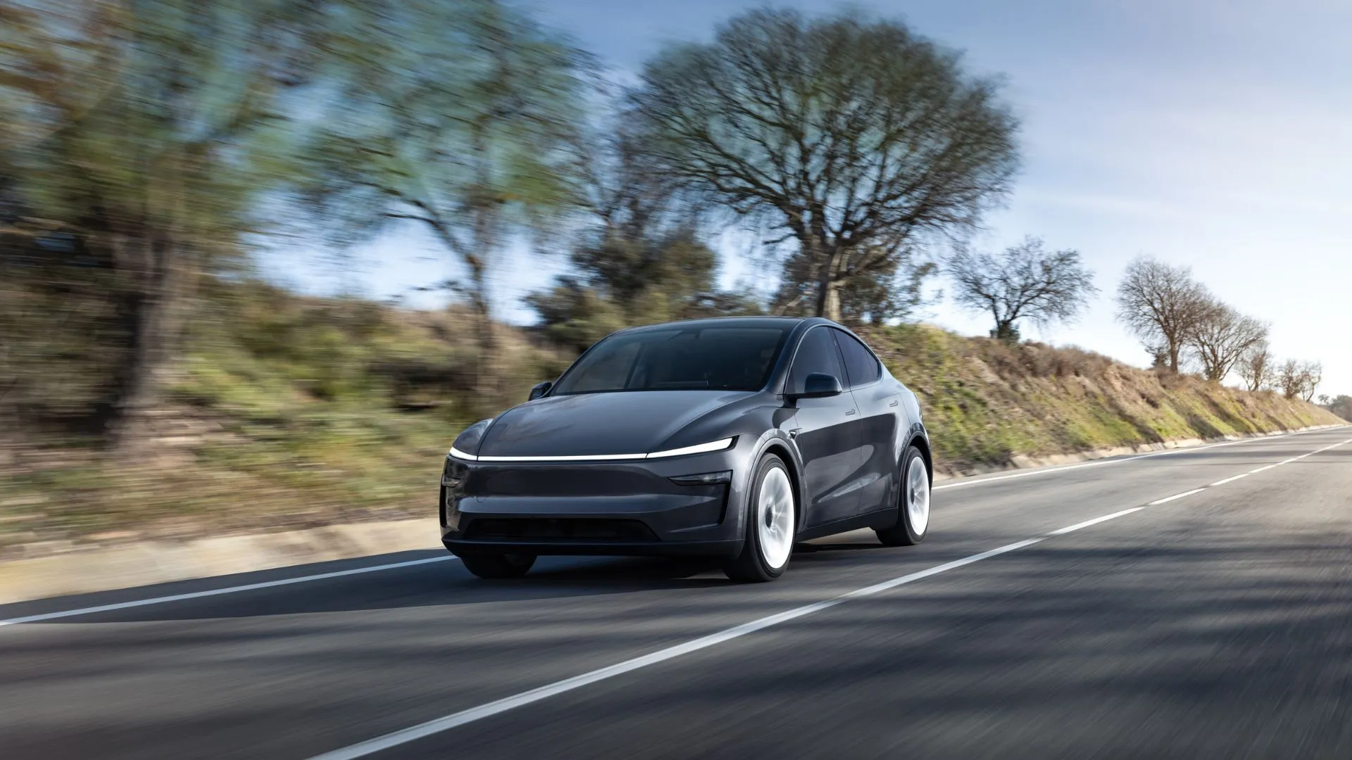 Mẫu SUV điện Tesla Model Y 2025 màu xám di chuyển trên đường nông thôn, thể hiện sự cải tiến về phong cách.