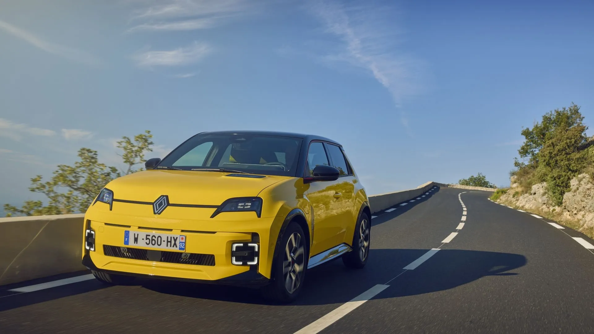 Mẫu xe điện Renault 5 E-Tech phiên bản màu vàng tươi tắn, đứng trước phông nền đô thị, nổi bật với thiết kế retro hiện đại và logo Renault phát sáng.