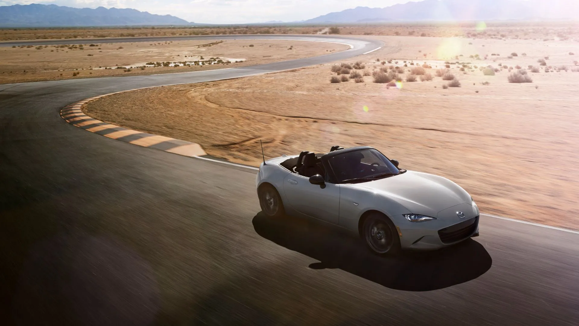 Mazda MX-5 Miata 2024 màu bạc đang di chuyển nhanh trên đường đua trong sa mạc, biểu tượng của sự nhẹ nhàng và tốc độ.