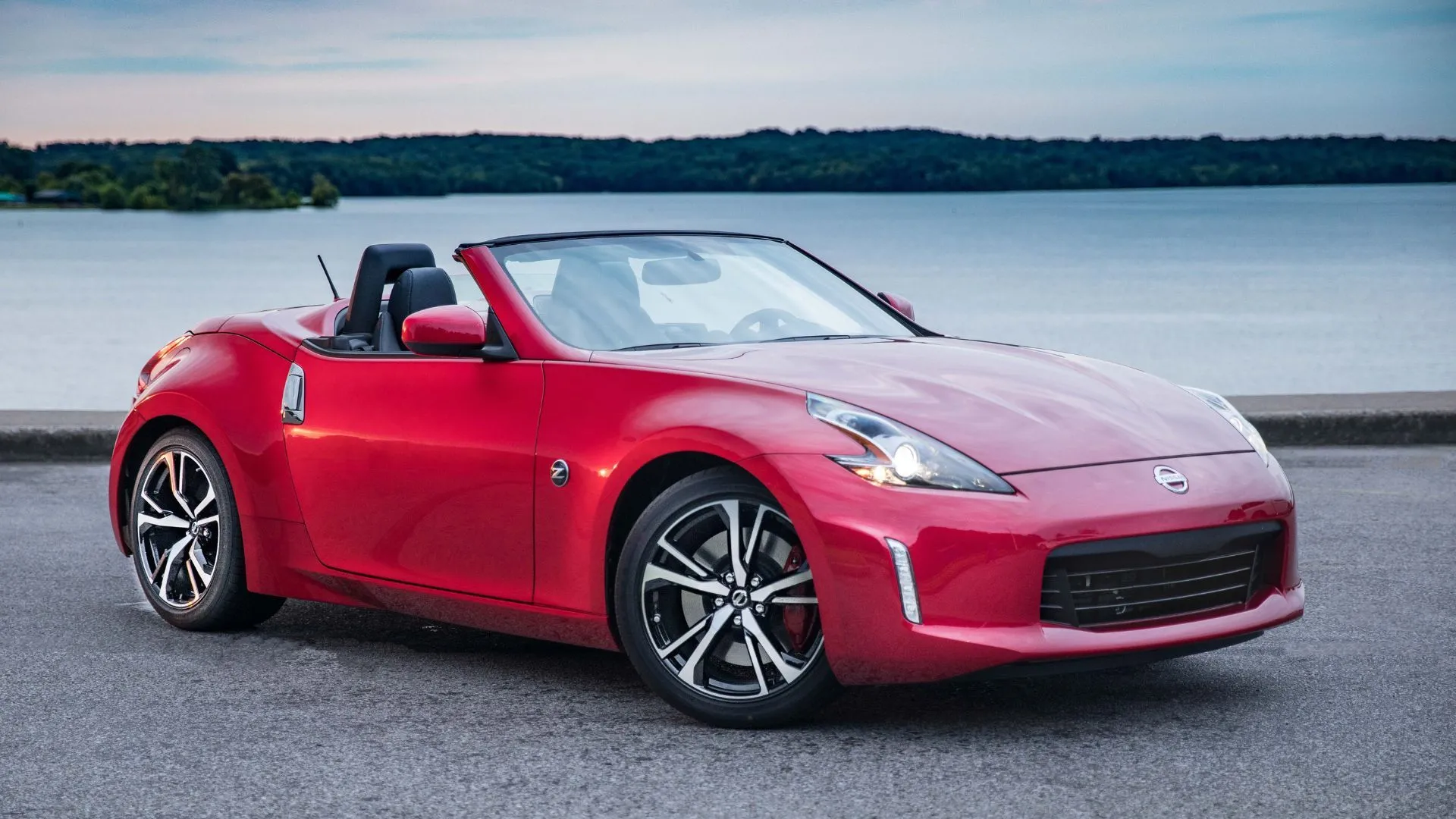Nissan 370Z Roadster 2019 màu đỏ quyến rũ với mui trần mở, đậu bên bờ nước, tạo nên hình ảnh thể thao và phong trần.