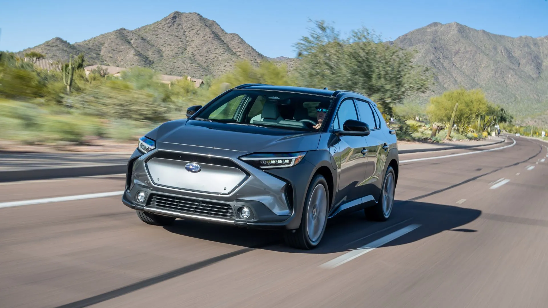 Subaru Solterra 2024 màu xám đang lướt trên con đường nông thôn, phía sau là những ngọn núi hùng vĩ, thể hiện khả năng off-road nhẹ của SUV điện.