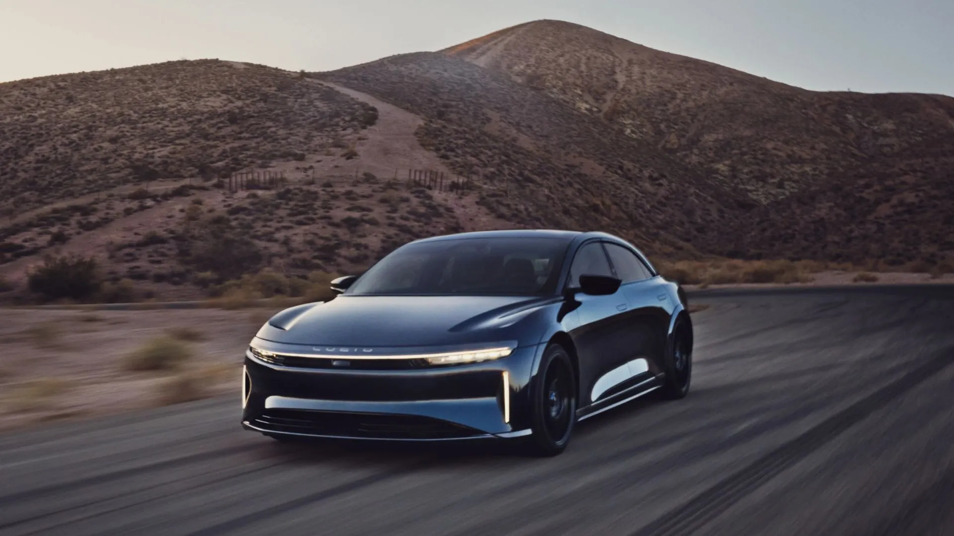 Toàn cảnh chiếc Lucid Air Sapphire màu xanh lam đang lái trên một con đường nông thôn với núi non ở hậu cảnh