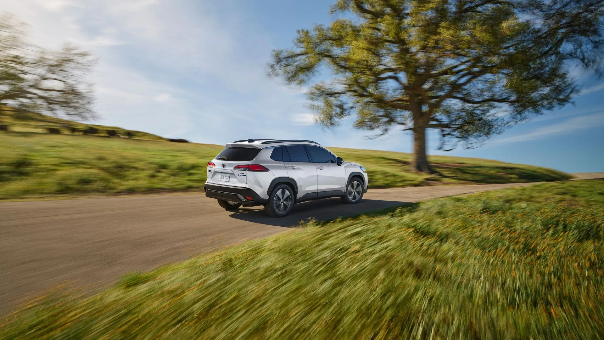 Toyota Corolla Cross Hybrid XSE 2025 màu trắng ngọc trai Wind Chill Pearl đang di chuyển trên con đường nông thôn quanh co với cây xanh, toát lên vẻ thanh lịch và linh hoạt