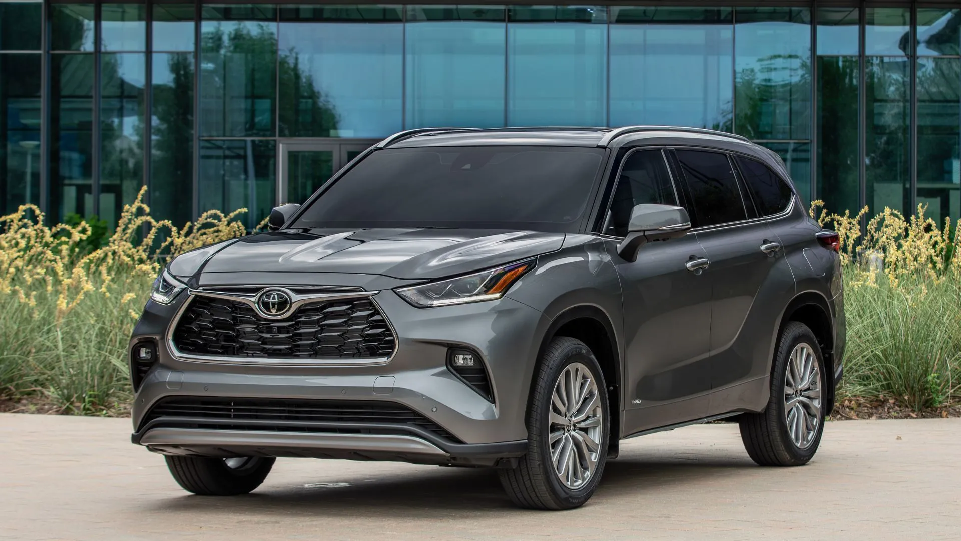 Toyota Highlander Hybrid 2025 màu xám đỗ trước tòa nhà kính, nổi bật với khả năng tiết kiệm nhiên liệu tối ưu.