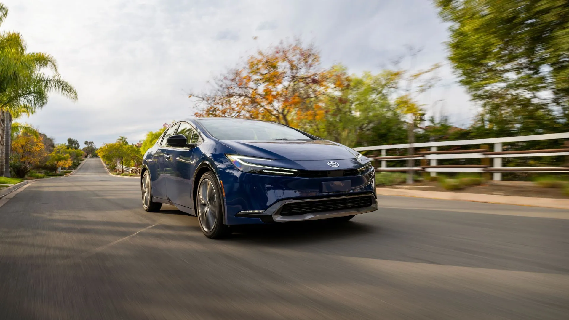 Toyota Prius LE 2025 màu xanh đậm sang trọng di chuyển trên con đường quê, minh chứng cho thiết kế hiện đại và khả năng tiết kiệm nhiên liệu tối ưu.
