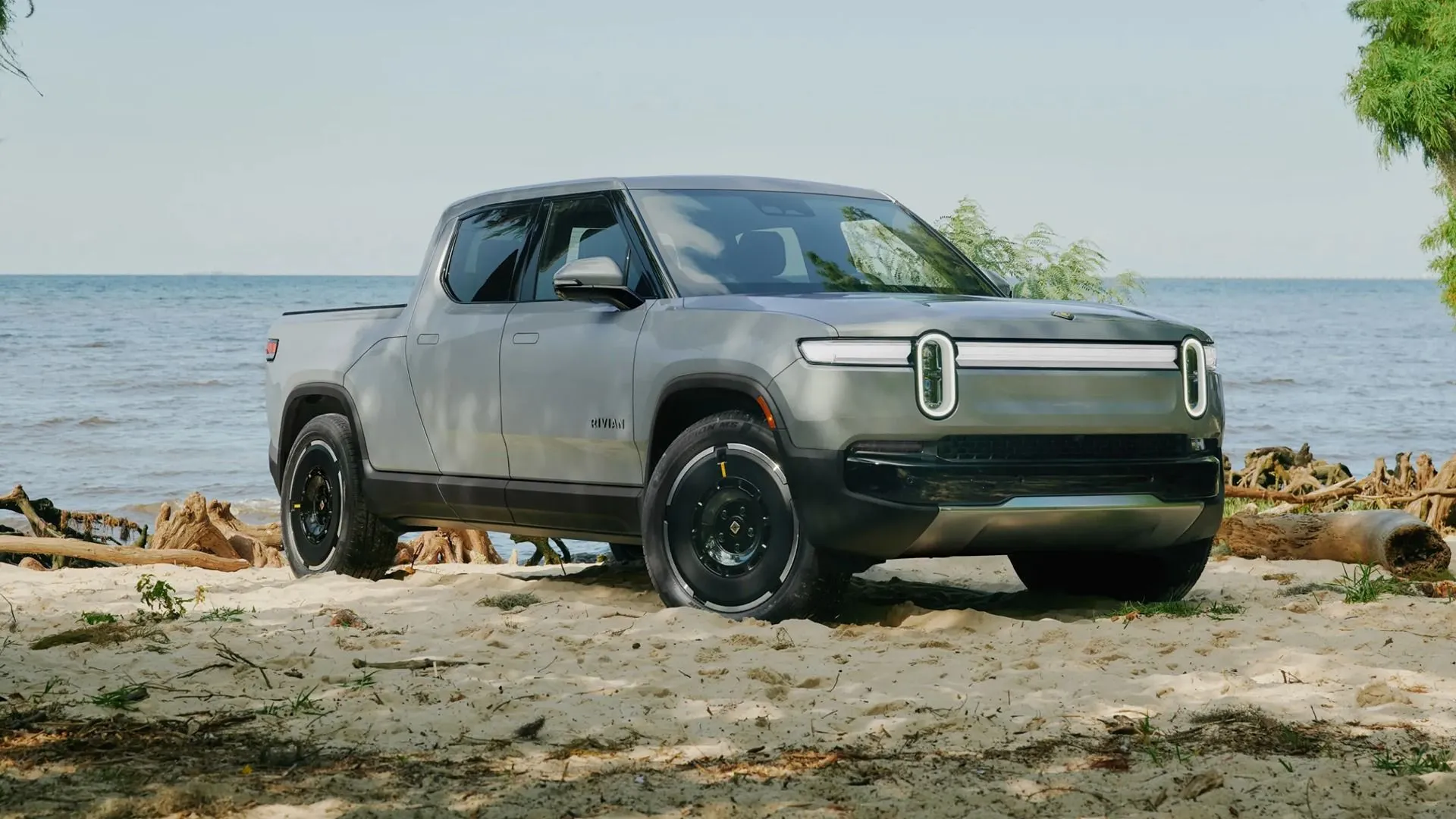 Xe bán tải điện Rivian R1T 2025 màu xám với góc nhìn phía trước trên bãi biển, cho thấy sự linh hoạt và sang trọng.