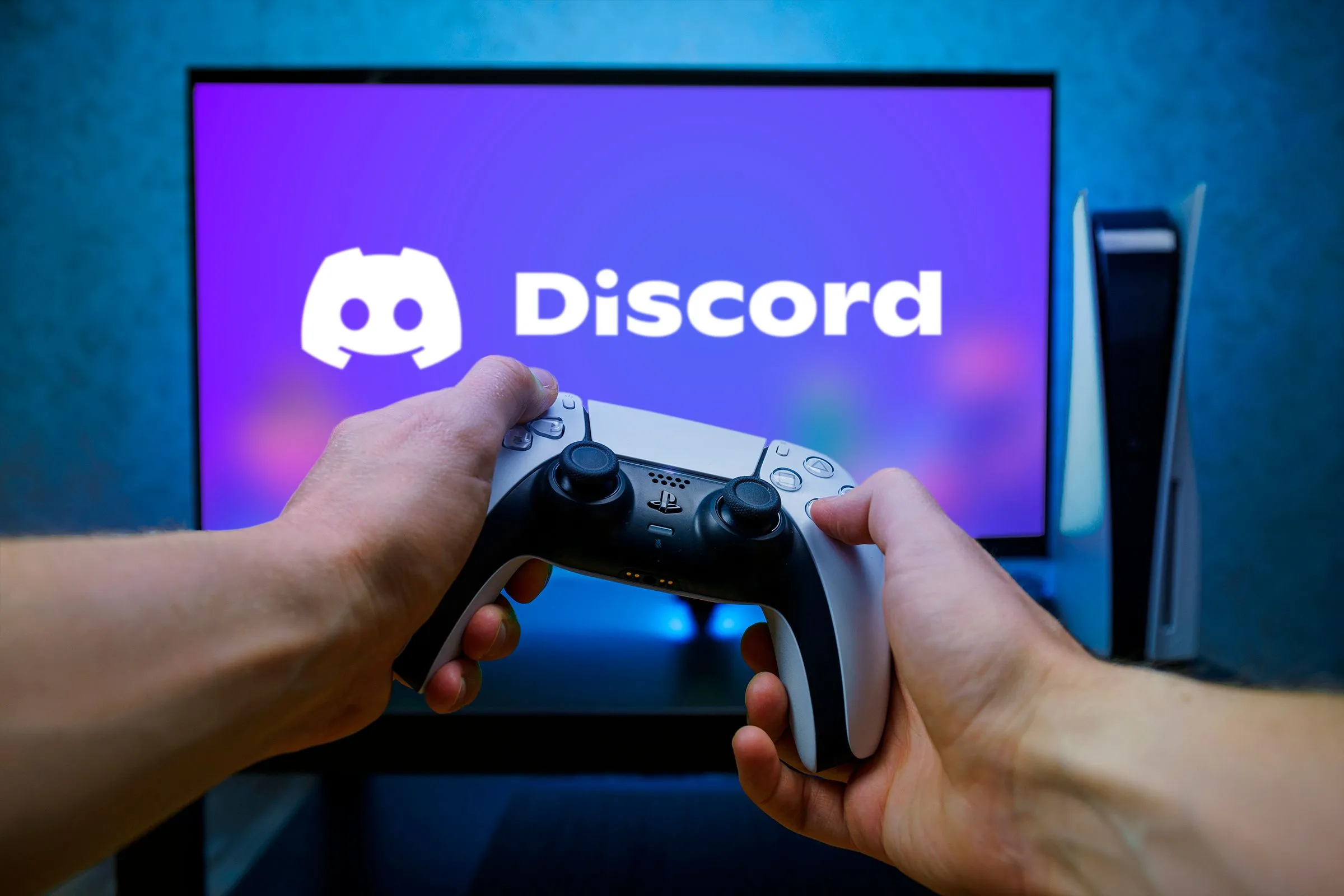 Một người đang cầm bộ điều khiển PS5 DualSense với màn hình TV phía trước hiển thị logo Discord, ứng dụng giao tiếp cộng đồng phổ biến